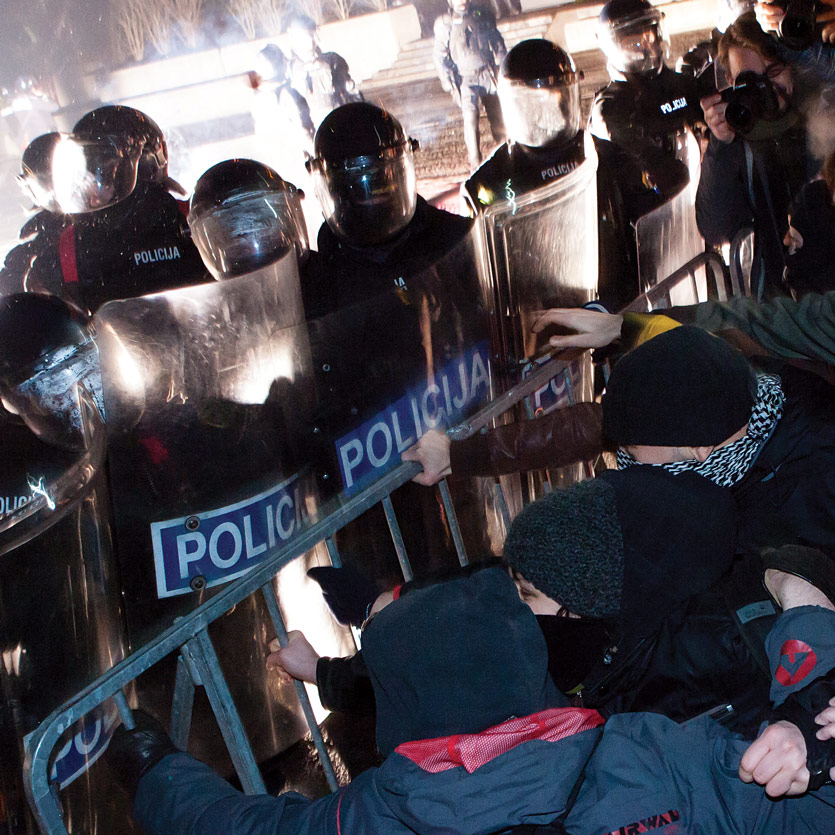 a mustachioed cop in riot gear wearing aviator sunglasses stands guard in front of several Ku Klux Klan members in full robes, one of whom carries a American flag